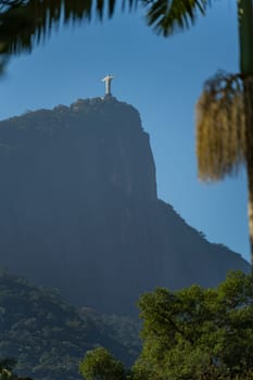 View of iconic Christ the Redeemer in Rio de Janeiro from below, framed by palm tree, overlooking a rocky cliff under clear blue sky.