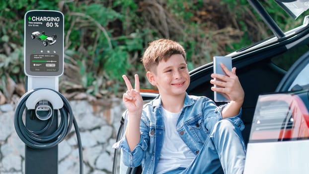 Little boy sitting on car trunk taking selfie using smartphone while recharging eco-friendly car from EV charging station. EV car road trip using sustainable energy concept. Perpetual