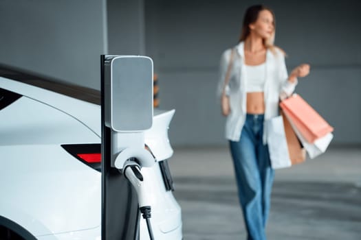 Young woman travel with EV electric car to shopping center parking lot charging in downtown city showing urban sustainability lifestyle by green clean rechargeable energy of electric vehicle innards