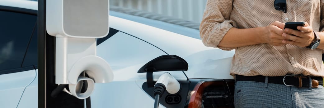 Young man use smartphone to pay for electricity at public EV car charging station at city commercial mall parking lot. Modern environmental and sustainable urban lifestyle with EV vehicle. Expedient