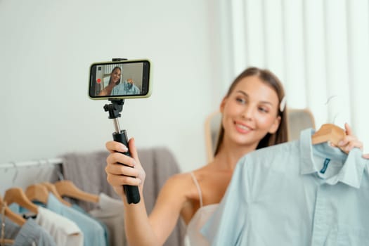 Young woman making fashion video content for social media. Blogger smiles to camera using selfie stick and light ring while making persuasive online clothing sell to audience or followers. Unveiling