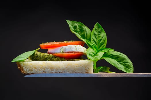 fresh slice of bread with pesto sauce, mozzarella and tomato on a wooden spatula with basil. Isolated on a black background.