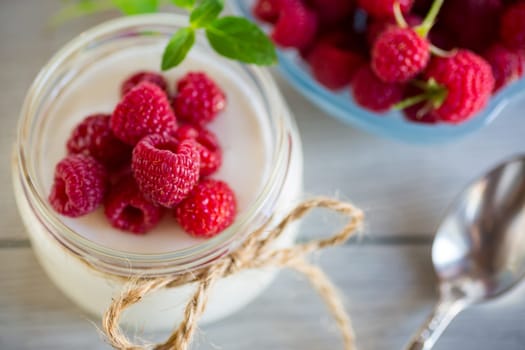 Sweet cooked homemade yogurt with fresh raspberries in a glass jar, on a light wooden table.