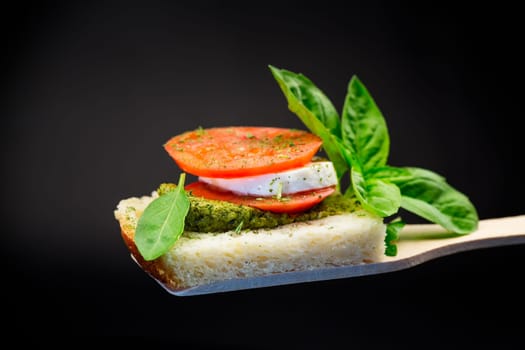 fresh slice of bread with pesto sauce, mozzarella and tomato on a wooden spatula with basil. Isolated on a black background.