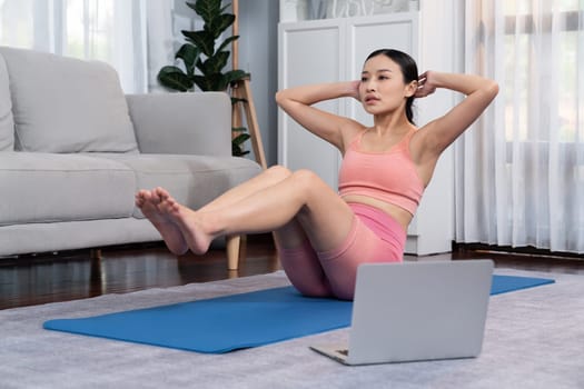 Asian woman in sportswear doing crunch on exercising mat as home workout training routine. Attractive girl engage in her pursuit of healthy lifestyle with online exercise training video. Vigorous