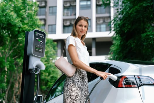 Young woman travel with EV electric car charging in green sustainable city outdoor garden in summer. Urban sustainability lifestyle by green clean rechargeable energy of electric BEV vehicle innards