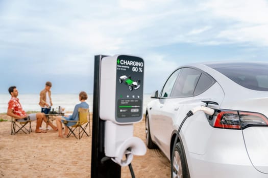 Alternative family vacation trip traveling by the beach with electric car recharging battery from EV charging station with blurred family enjoying the seascape background. Perpetual