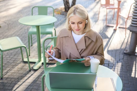 Portrait of young blond woman, female student in street cafe, wearing wireless headphones, using laptop, having online meeting, attend web lecture or course, doing homework.