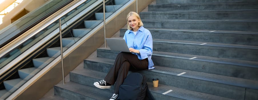 Portrait of young woman, student sitting outdoors on stairs with coffee, using laptop, working on project, looking at camera and smiling.