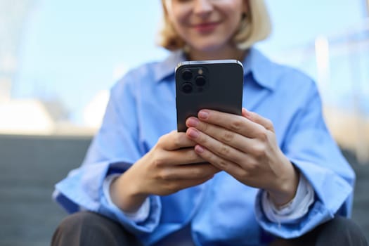 Close up portrait of young woman, holding smartphone in both hands, sitting on street with mobile phone, messaging and chatting on telephone.