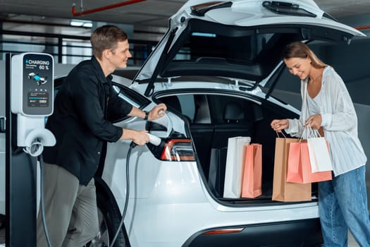 Young couple travel with EV electric car to shopping center parking lot charging in downtown city showing urban sustainability lifestyle by green clean rechargeable energy of electric vehicle innards