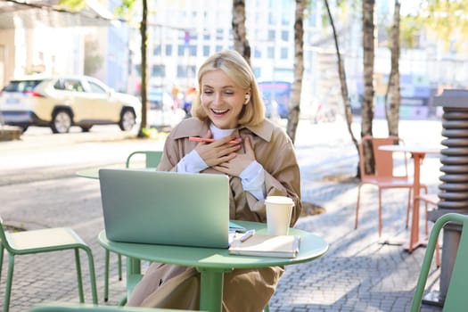 Portrait of young blond woman with laptop, sitting on street, drinking coffee in outdoor cafe, working, wearing wireless headphones, doing homework, freelancing.