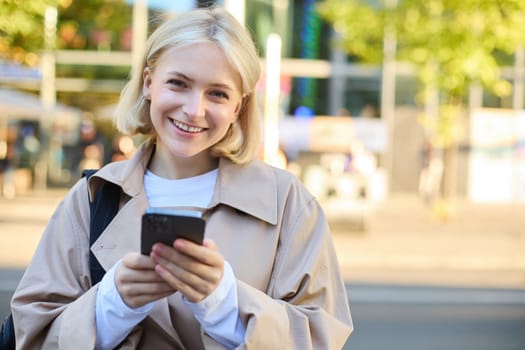 Close up portrait of young smiling blond woman, student on street, using mobile phone, checking messages on smartphone, walking on road.