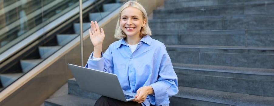 Image of smiling young woman, sitting on stairs outside university or college, waving hand, saying hello to passer-by, working on laptop.