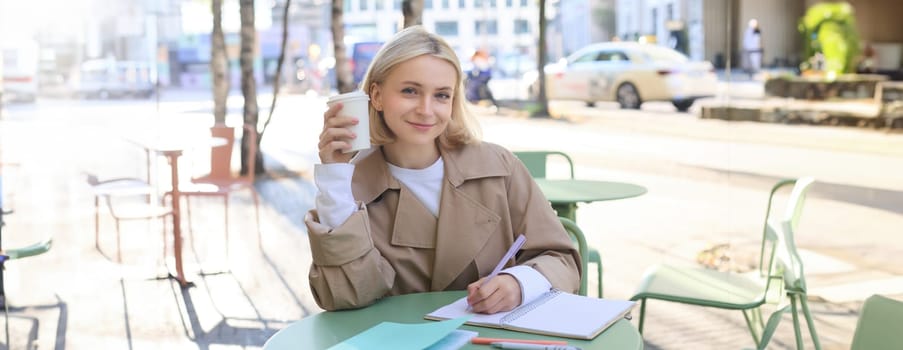 Image of young smiling woman, female entrepreneur writing, making corrections in documents, using notebook, drinking coffee and sitting in outdoor cafe.