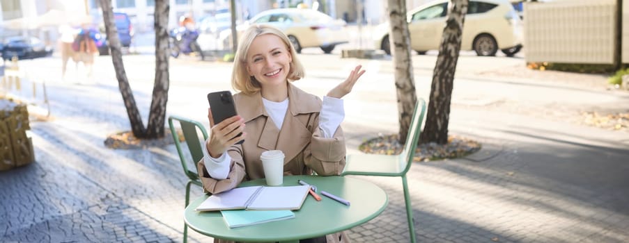 Happy young blond female model in coffee shop, talks on video chat, using smartphone and having conversation while resting outdoors in city centre.