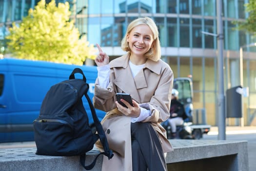 Portrait of young happy female student, woman sits on bench with backpack and phone, pointing finger at upper left corner, showing advertisement or banner.