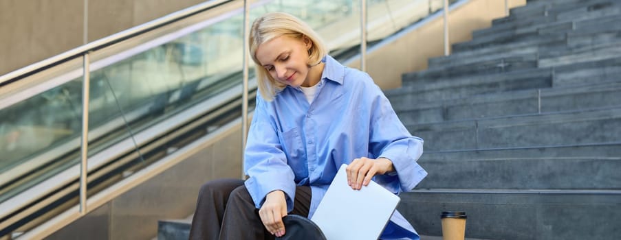 Smiling young female model, packing her laptop in backpack, sitting on city stairs outdoors, drinking cup of coffee before lecture in university.