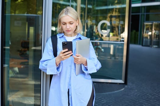 Image of young blond woman in blue shirt, holding laptop, waiting for someone near office building, using smartphone, mobile phone application.