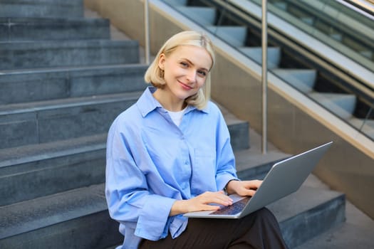 Portrait of woman with laptop, found wifi spot, sitting on stairs outdoors, drinking coffee, working on computer.