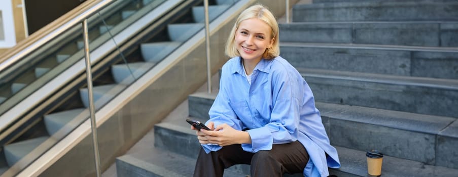 Lifestyle portrait of young urban female model, student drinks cup of takeaway coffee, sitting on stairs outdoors, using mobile phone, smiling and looking happy while taking a break.