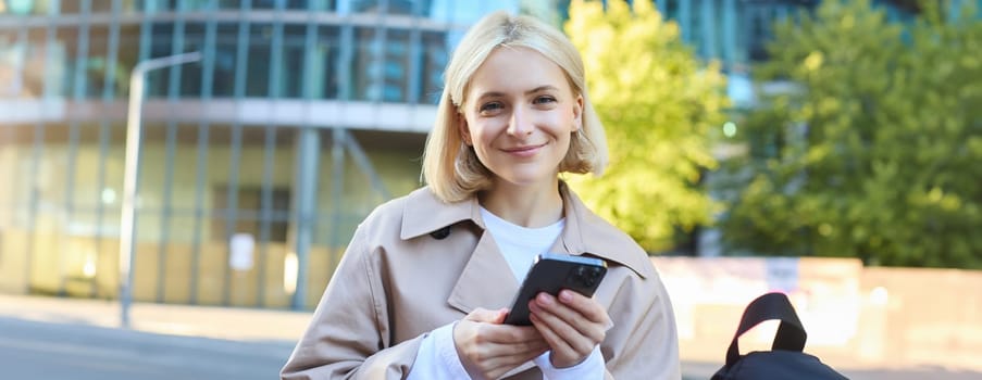 Close up portrait of happy blond girl waiting outside, standing on street with smartphone, looking at camera and smiling.