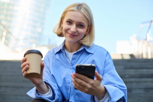 Portrait of beautiful blond woman, smiling girl student drinking coffee, sitting on stairs with smartphone, looking happy at camera.
