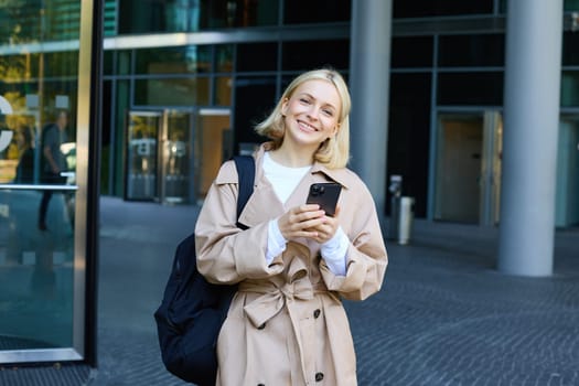 Portrait of young stylish woman walking in city centre with smartphone, using mobile phone app outdoors, waiting for someone, going to work.