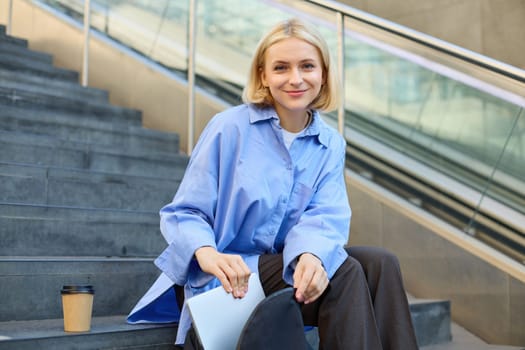 Young woman sitting on street stairs, packing her things, laptop inside backpack pocket, getting ready to go, spending time outdoors.