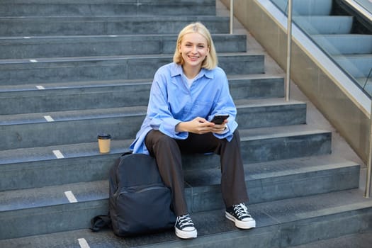 Portrait of woman on stairs, sitting with backpack, using mobile phone, messaging, scrolling social media on lunch break in university.