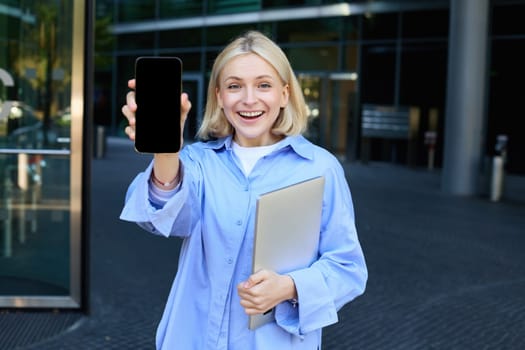 Happy smiling woman showing smartphone screen, demonstrating mobile phone display, standing in college outdoors, posing near campus with laptop and telephone.