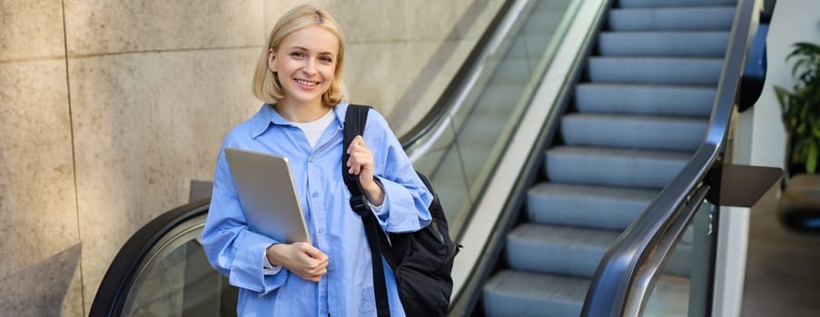 Concept of education and people. Young woman with backpack, carries laptop in hand, standing near escalator, going to tube, on her way to work on university.