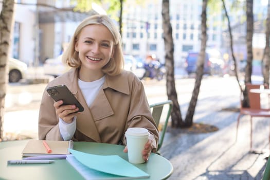 Portrait of young modern woman, student having her morning cup of coffee in city cafe, sitting outdoors, using mobile phone at the table, smiling at camera.