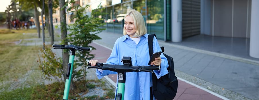 Portrait of young woman, renting a scooter, using mobile phone app to unlock it, using quick ride to get to work, smiling and looking happy.