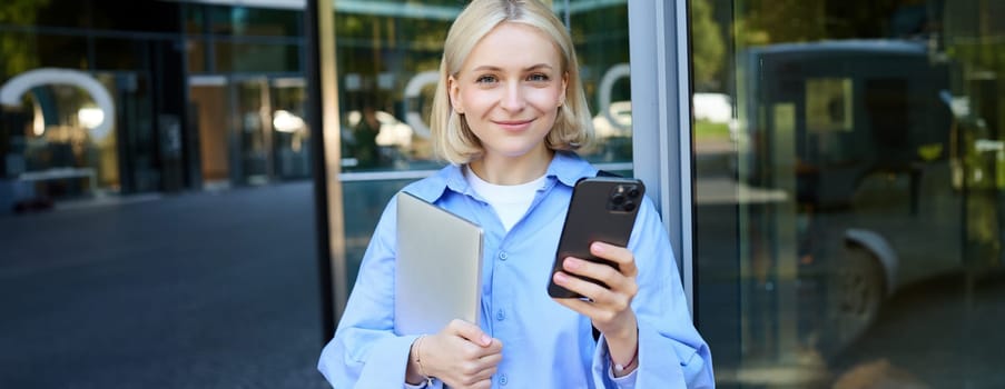 Close up portrait of young female student, blond girl with laptop and smartphone, using mobile phone app on street, standing near office building.