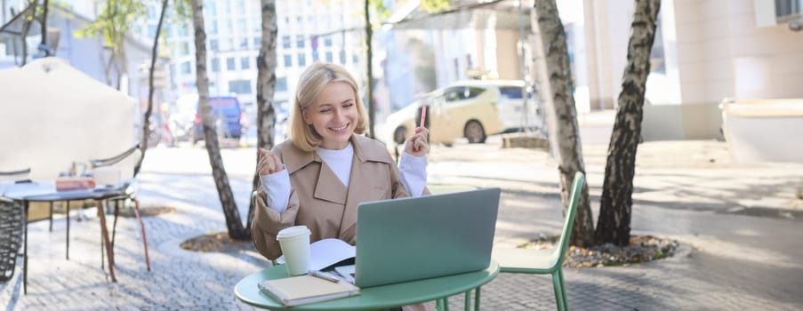 Remote workplace. Smiling young woman freelancer, student using laptop in outdoor cafe, talking to someone, attends online lecture, drinking coffee.