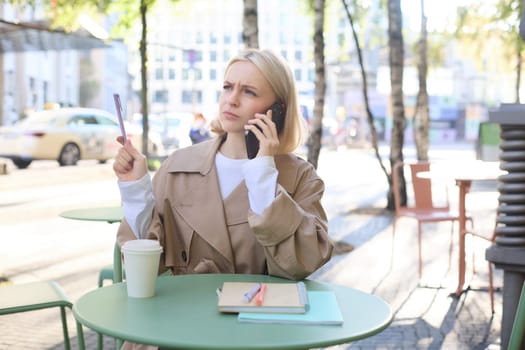 Image of woman with confused, serious face, answers phone call, working in cafe outdoors, drinking coffee, frowning with pen in hand.