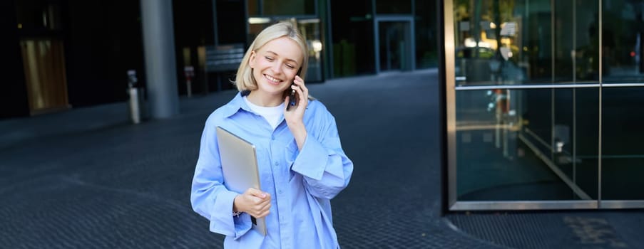 Portrait of young businesswoman in blue collar shirt, talking on mobile phone, standing near office building and smiling, holding work laptop.