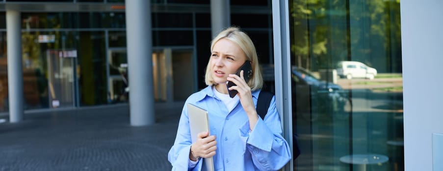 Portrait of young modern woman, office manager near building, standing outside with backpack, laptop, talking on mobile phone, chatting on smartphone with confused face.