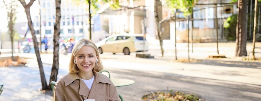 Vertical portrait of smiling blond woman, female student with coffee and laptop, has notebooks on table, studying remotely, working for online project, spending time outdoors on sunny day.