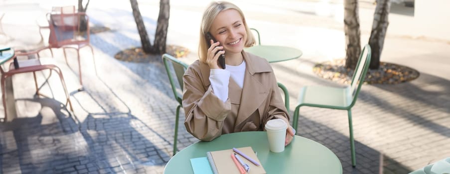 Image of stylish blond woman talking on mobile phone, drinking coffee in cafe outdoors, enjoying warm weather in city centre, answer telephone call on smartphone.