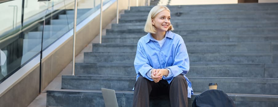 Portrait of young urban woman, college student with backpack, laptop and coffee, sitting on stairs in city, resting outdoors, taking break.