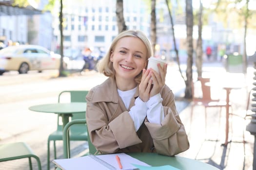 Happy young European woman, drinking coffee, sitting in street cafe, working and chatting, smiling at camera.