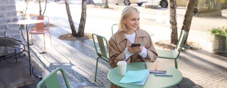 Portrait of blond smiling woman sitting in cafe, enjoying cup of takeaway coffee, using mobile phone, working on smartphone, social media blogger making post online.