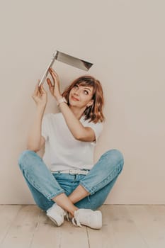 A brunette sits on the floor with a laptop on a beige wall background. She is wearing a white T-shirt, jeans and white sneakers