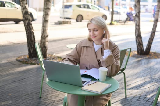 Young woman, freelancer, student doing homework in outdoor cafe, drinking her coffee on street, using laptop, connects to online lecture or doing course in internet.