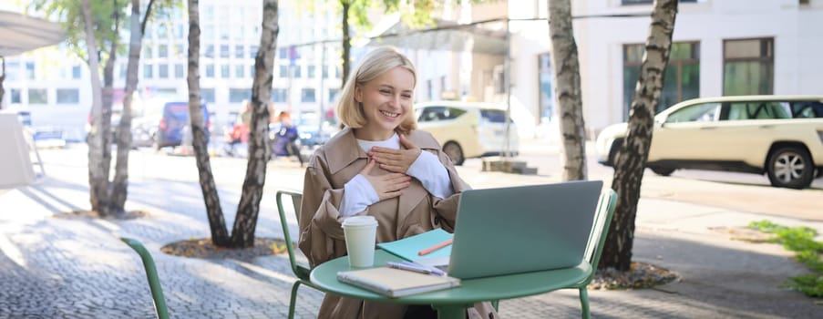 Image of young smiling woman working remotely, connects to online call, chatting via laptop, looking pleased, feeling grateful, laughing, sitting in outdoor cafe.