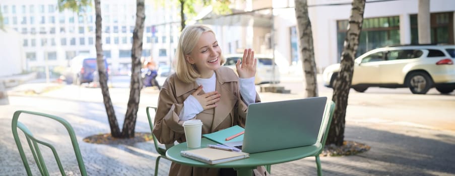 Portrait of young friendly woman, waving hand at laptop and smiling, saying hello, greeting someone, online chatting, having remote conversation while sitting in coffee shop.