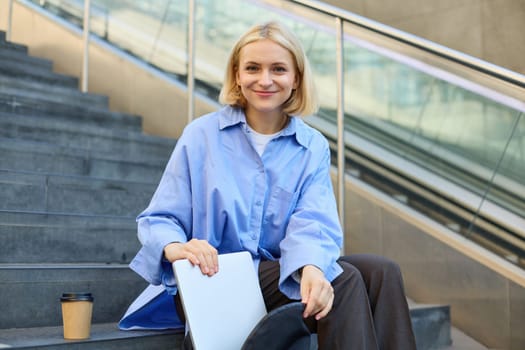 Portrait of beautiful smiling woman, student on stairs in city, sitting and drinking coffee, packing laptop in backpack, looking happy at camera.