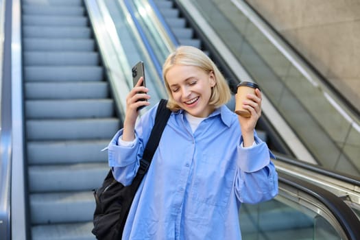 Image of young happy blond woman, dancing from happiness and joy, drinking takeaway coffee, holding smartphone, posing near escalator in city centre.